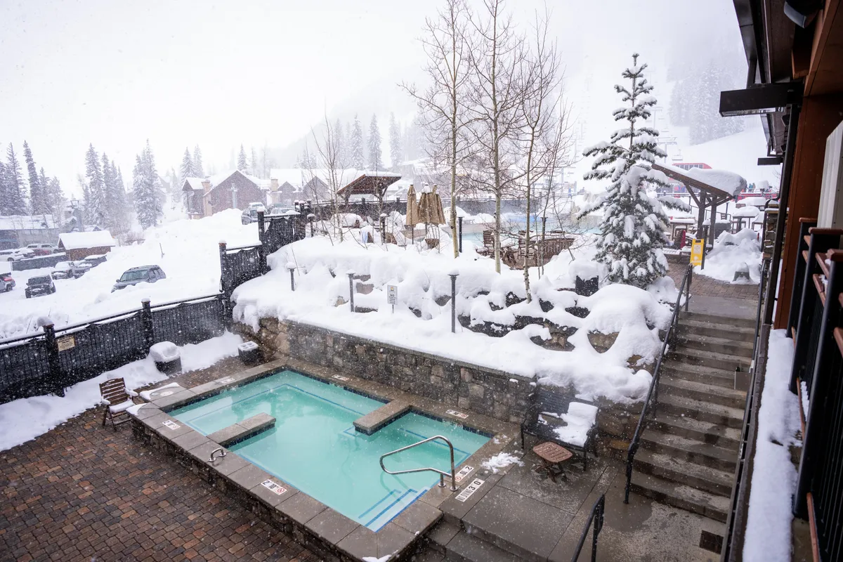 A picturesque outdoor rooftop hot tub set against a snowy landscape, with steps leading up to the steaming water. People are relaxing in the hot tub, enjoying the tranquil ambiance and mountain scenery as snow gently falls around them. The scene evokes a sense of warmth, relaxation, and social connection.