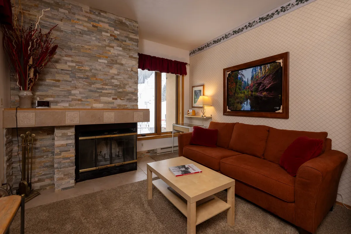 A warmly lit lounge area featuring a classic wood-burning fireplace, a plush sofa, and a coffee table, with artistic decor on the walls. The space feels inviting, perfect for gathering, relaxing, or enjoying quiet moments after outdoor adventures.