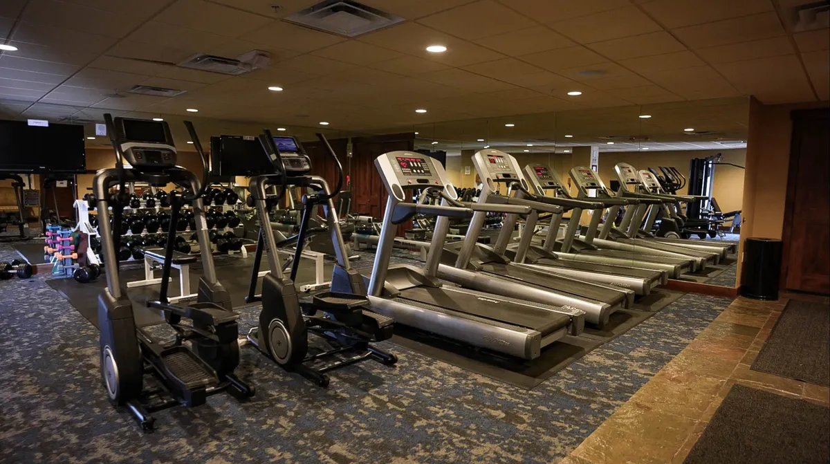 A modern, well-equipped fitness center filled with treadmills, ellipticals, weightlifting equipment, and large mirrors. Several people are engaged in workouts, group classes, or personal training sessions, emphasizing an energetic and motivational environment.
