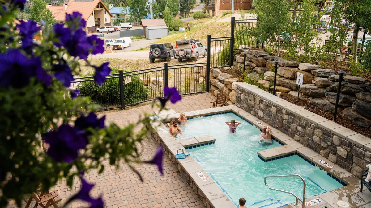A lively outdoor swimming pool surrounded by colorful flowers and a mix of guests sunbathing, swimming, and socializing. The pool area is vibrant, with people enjoying the water and the leisurely atmosphere under the open sky, suggesting an inviting and inclusive resort experience.