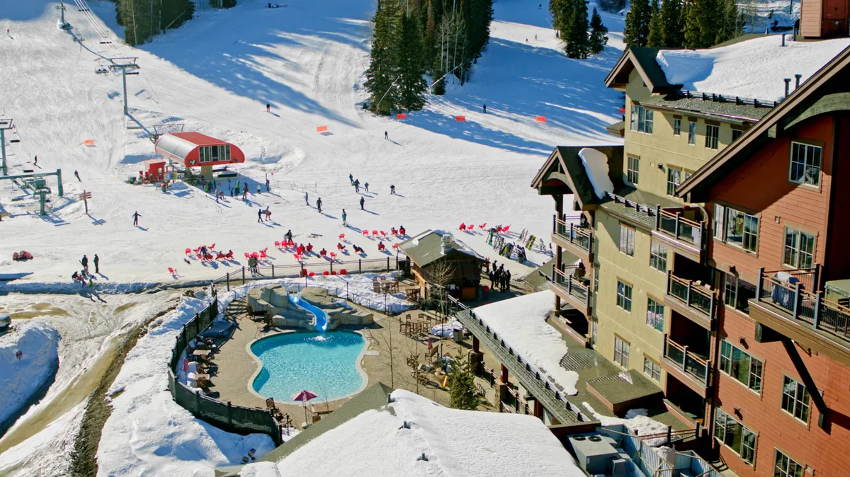 This energetic exterior shot captures the ski slopes bustling with activity, a swimming pool, the building’s exterior, a ski lift, and people enjoying various outdoor pursuits including skiing and outdoor dining.