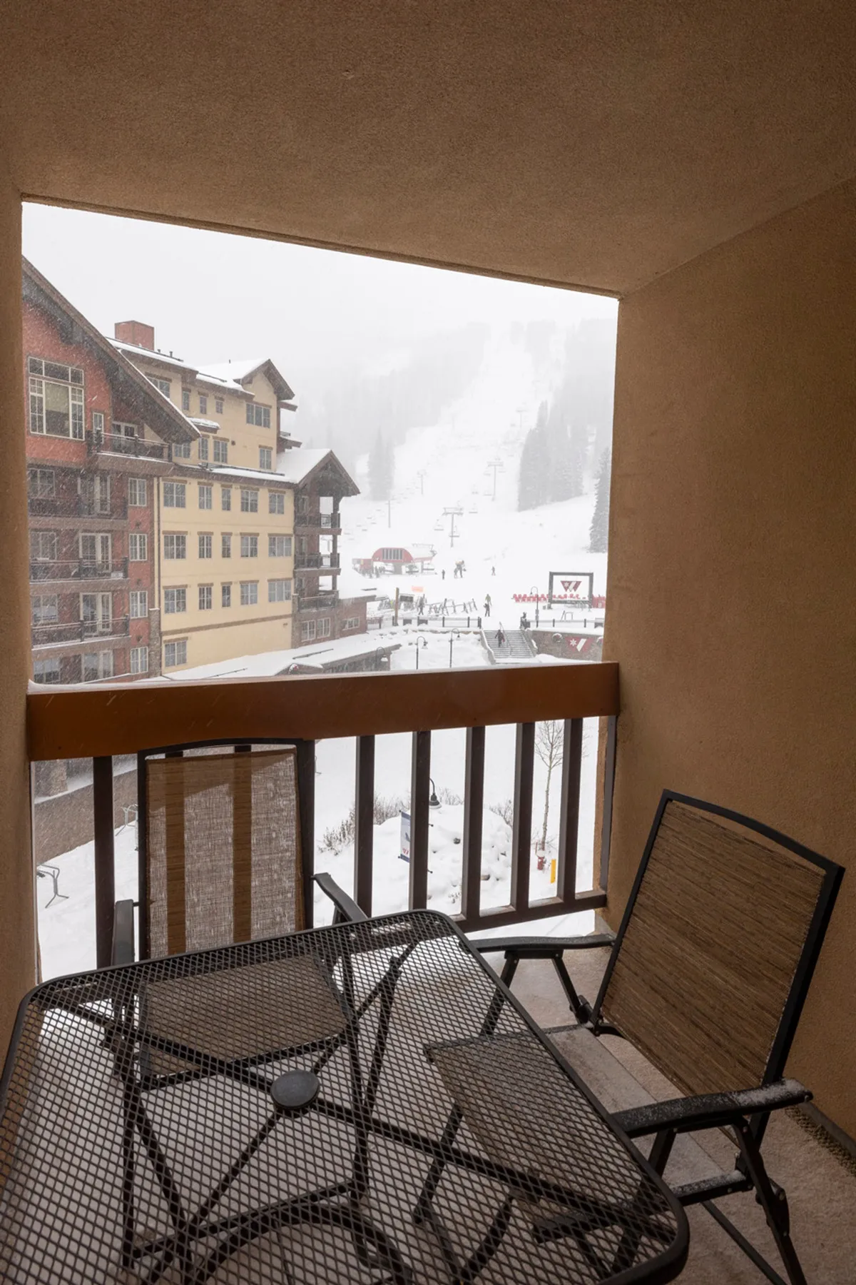 This image features a snow-covered balcony with a table and chairs, offering a serene view of ski slopes and the surrounding buildings. The scene is inviting, suggesting a perfect spot to relax after skiing or enjoy a warm beverage while watching the action in the village below.