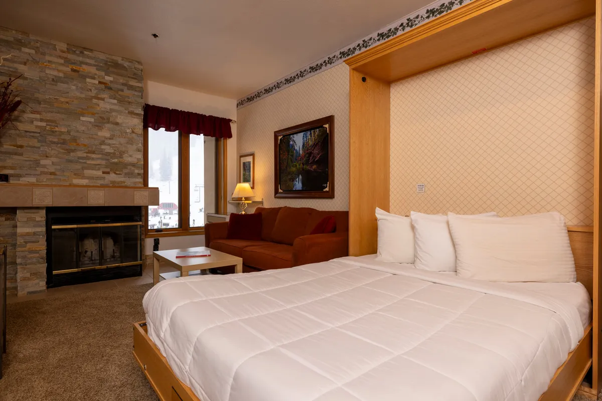 A cozy, open-plan studio interior featuring a plush bed nestled close to a glowing wood-burning fireplace. A comfortable sofa sits nearby, with large windows in the background letting in natural light and offering glimpses of mountain scenery. The layout feels intimate, with inviting textures and warm tones throughout, perfectly suited for relaxing after a day outdoors.