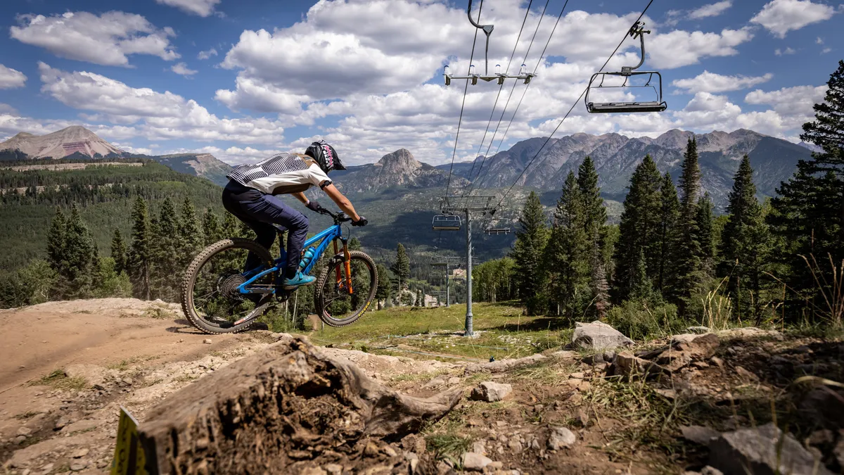 A vibrant summer mountain scene with a mountain biker riding a trail, a scenic chairlift in the background, expansive mountain ranges, and lush trees. The image radiates excitement and the spirit of outdoor exploration beyond winter.
