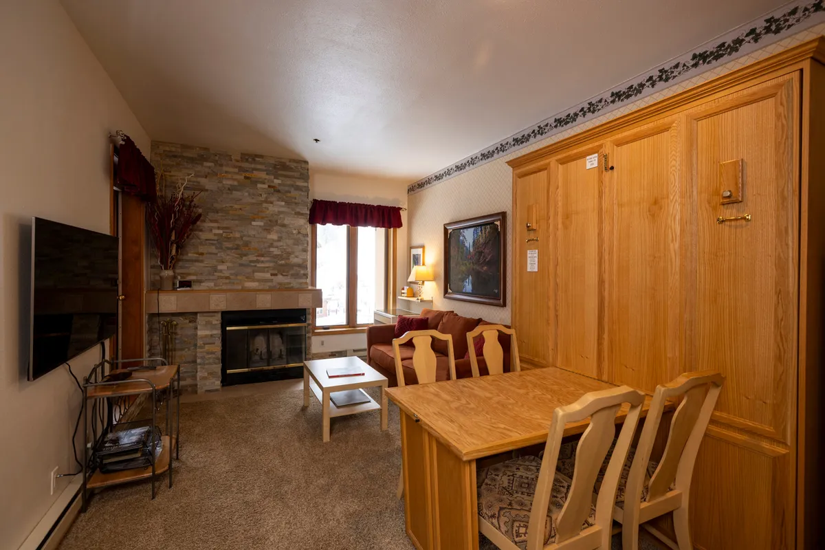 The image showcases a warmly lit studio interior with a crackling wood-burning fireplace as the focal point, a cozy seating area perfect for relaxing, a small dining table ideal for morning coffee or evening meals, and tasteful artwork adorning the walls. The setting exudes comfort and invites guests to unwind after a day on the nearby slopes.