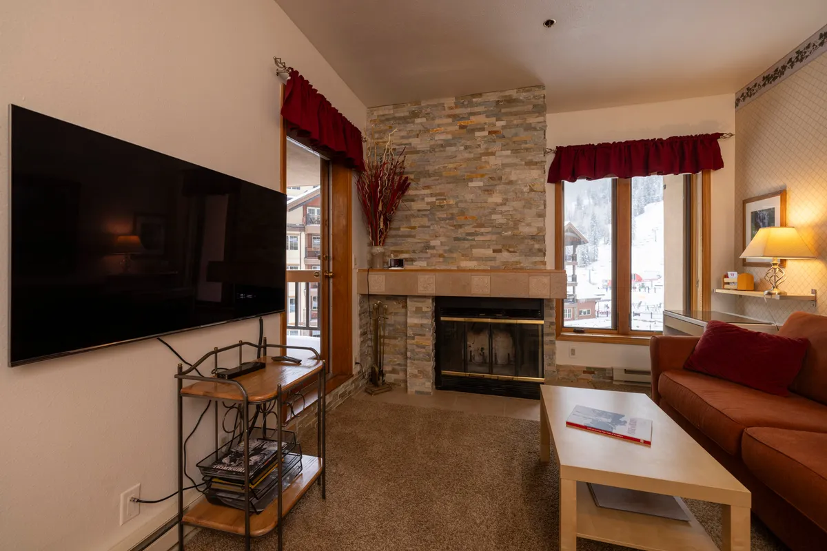 A bright, inviting living space centered around a fireplace and a soft, spacious sofa. A television and large windows are visible, with sunlight streaming in and highlighting the mountain or snowy landscape outside. The scene suggests a perfect spot for both relaxing evenings and bright, sun-filled mornings.