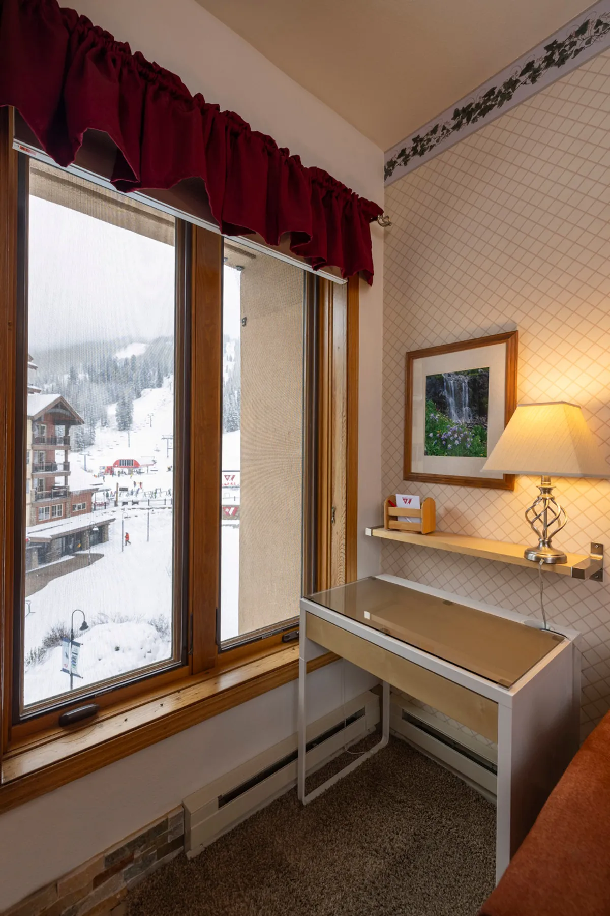 A peaceful nook beside large windows framing a snowy mountain view. A desk with a lamp sits under the window, inviting moments of reading, working, or simply enjoying the tranquil winter scenery. The scene is warm and thoughtfully designed, ideal for quiet relaxation.